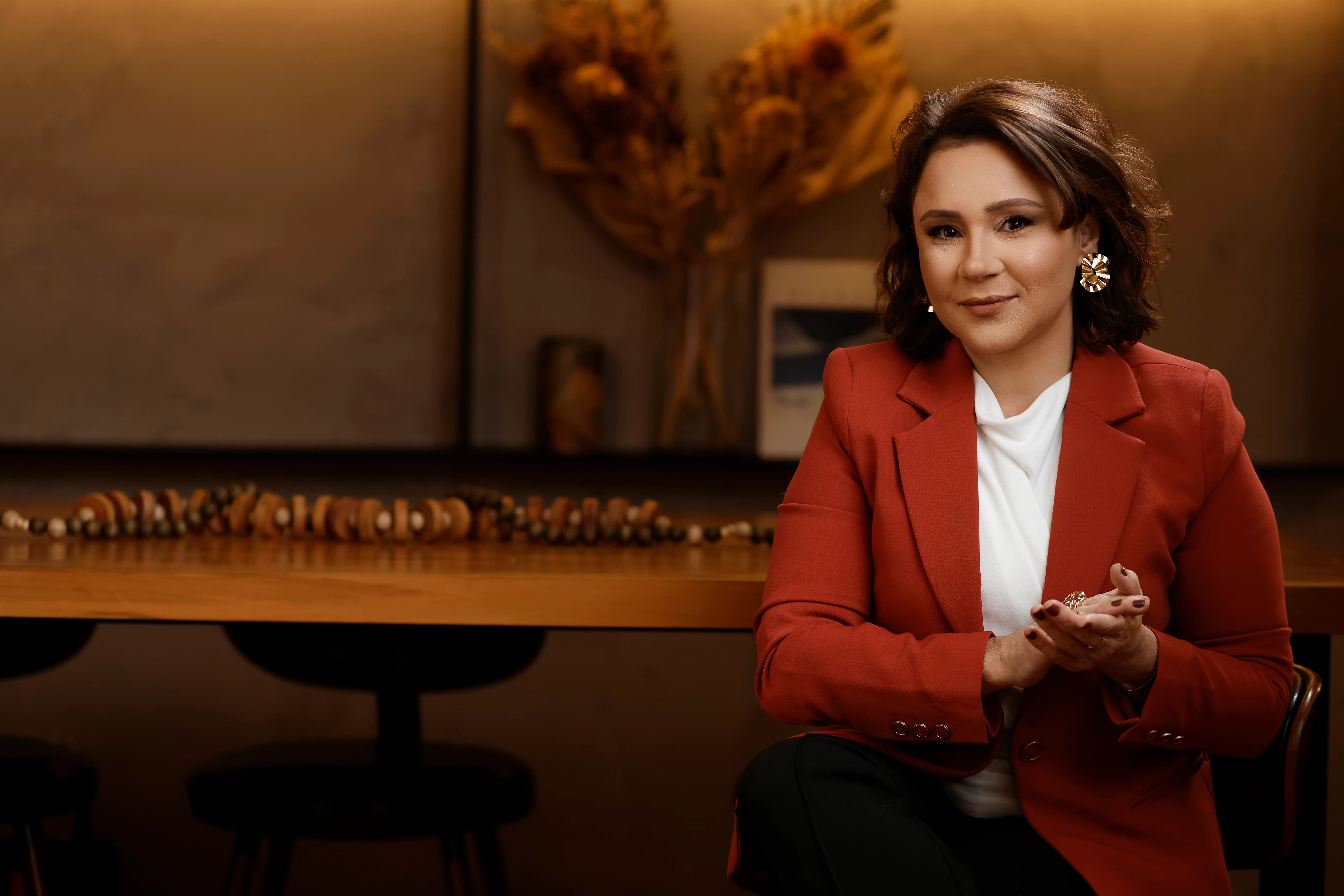 Katiuscia Silva, de blazer terracota, sentada com as mãos entrelaçadas e meio sorriso.