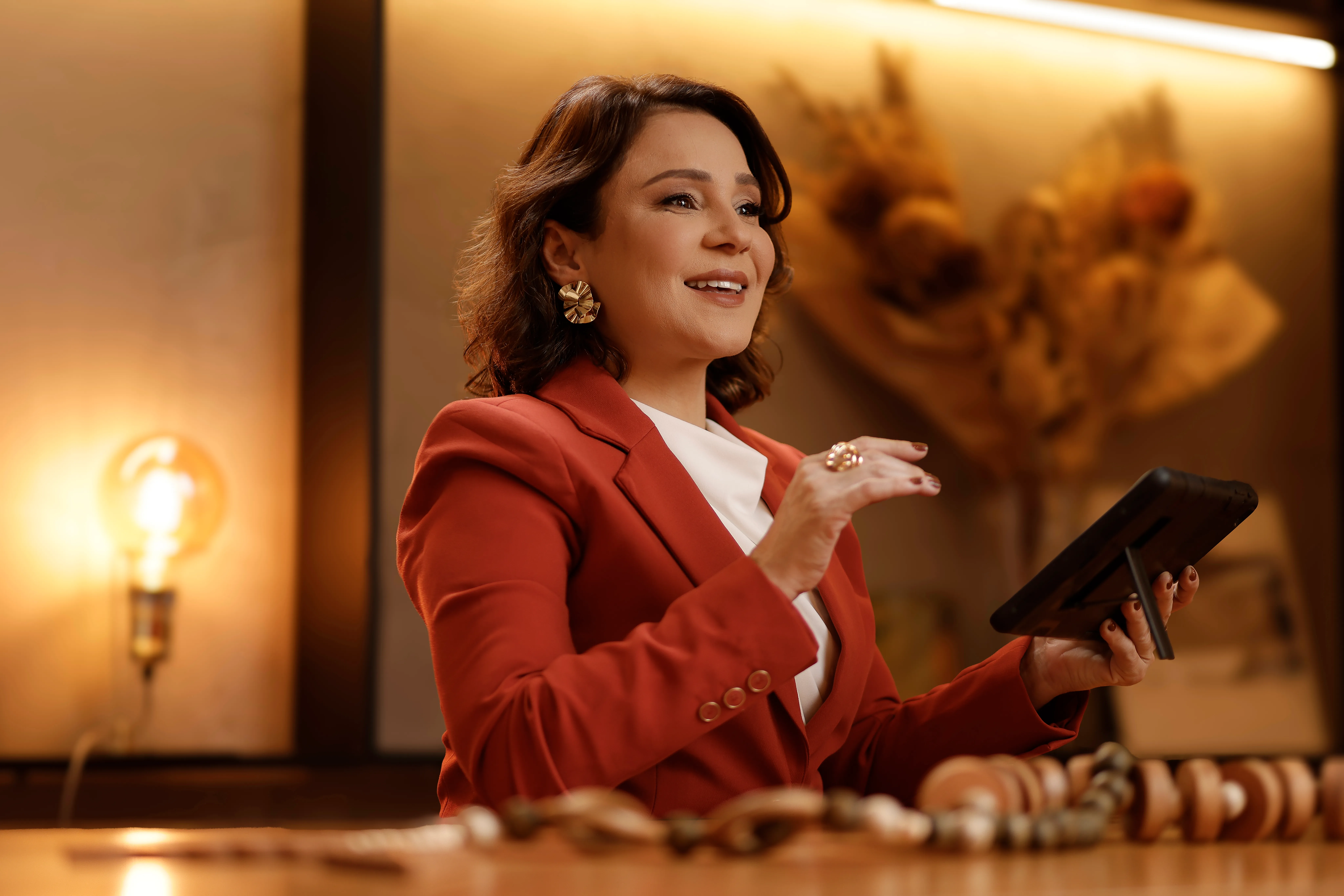 Katiuscia Silva, de blazer terracota, segurando um tablet e gesticulando durante uma explicação.